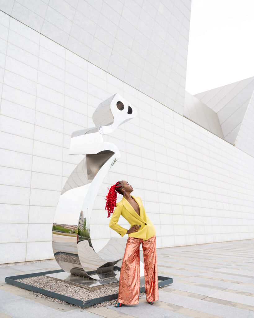 A black woman with curly red hair in a ponytail, posing with an abstract sculpture