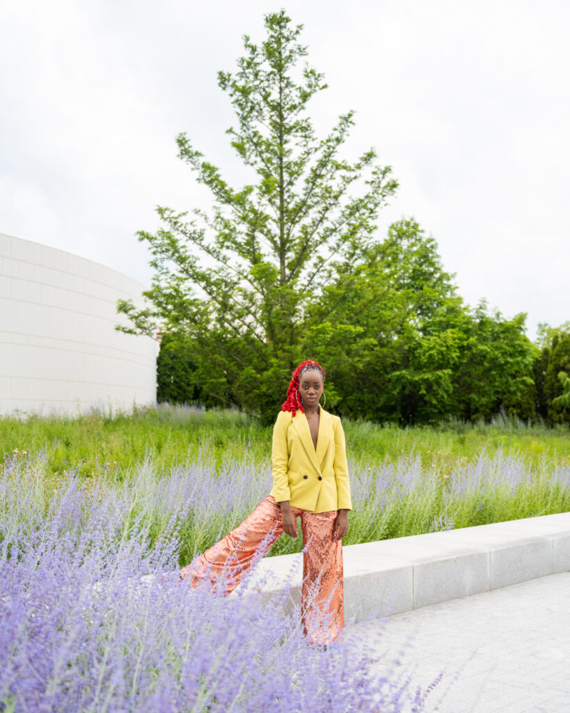 A black woman wearing a red ponytail, posing among lavender plants and greenery