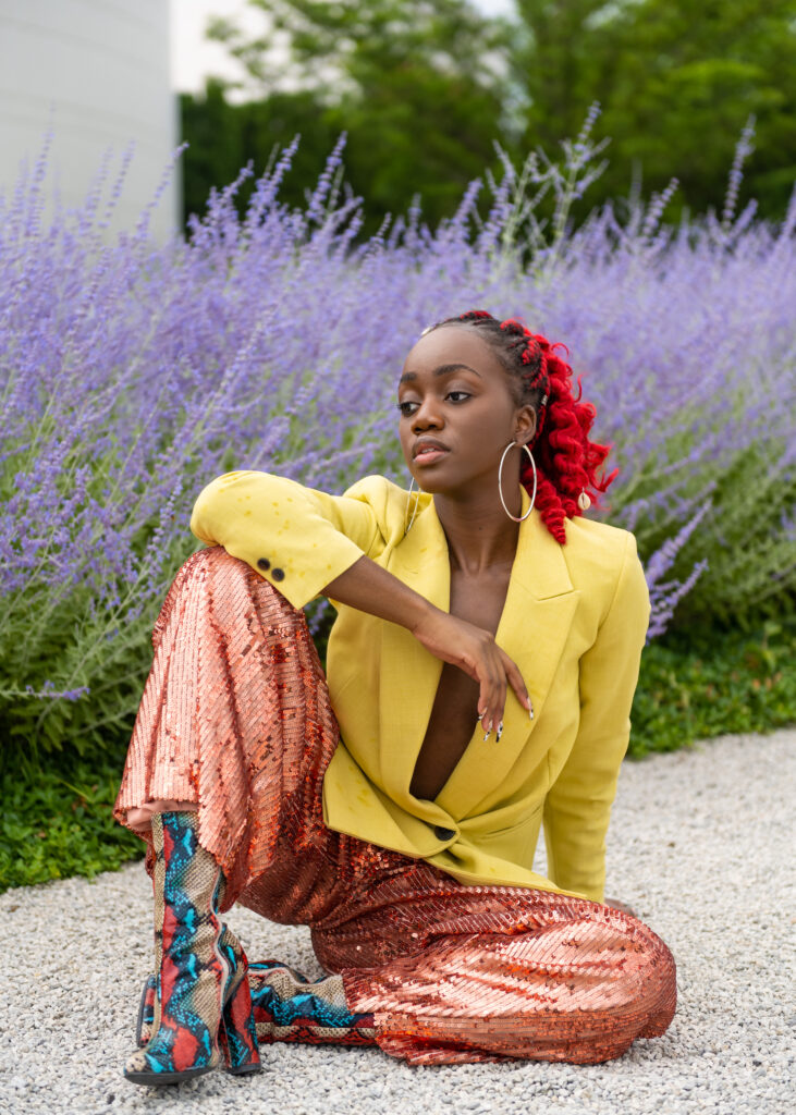 A black woman in a red ponytail, sitting on pebbled ground in front of lavender plants