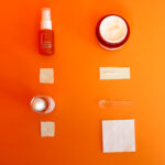 Tonal flat lay photograph of 3 skincare products and an orange against a solid orange coloured background.