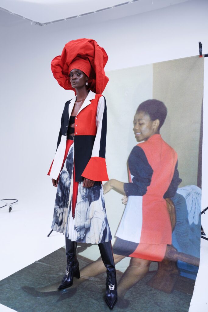 A tall Black person is staring directly into the camera with a stoic expression. They are wearing a bright red headdress, small silver earrings that resemble South African coins, a red, black, and white colour blocked blazer with flared sleeves, and a knee length pleated skirt with a black and white photograph printed on it. The model is wearing shiny, heeled, black boots that have Thebe Magugu's logo attached as a silver pendent. Behind the model is a backdrop that features a photograph of a Black person with low cut hair wearing a black, white, and red colour blocked blazer. Their back is turned and they are smiling while looking over one shoulder with their eyes lowered.